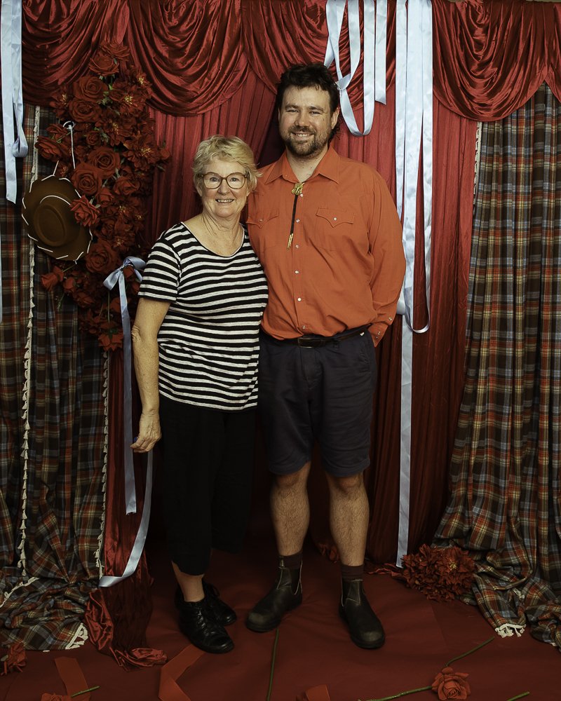 A smiling older woman and a smiling man stand together in front of red curtains with roses and plaid curtains, some blue ribbons, and a cowboy hat hanging on the left side.