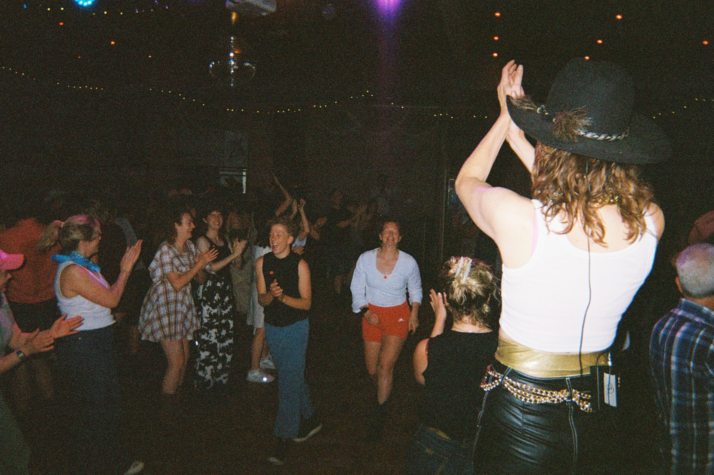 People dancing and enjoying music in a dark club or party setting, with one woman in the foreground wearing a large hat, sleeveless top, and leather pants with a chain belt.