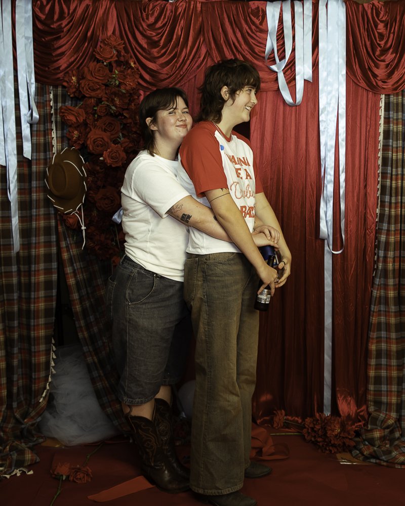 Two people standing in front of a red curtain backdrop with roses and plaid fabric, smiling and embracing. The person on the left wears a white t-shirt, shorts, and cowboy boots, while the person on the right wears a red and white t-shirt and brown p