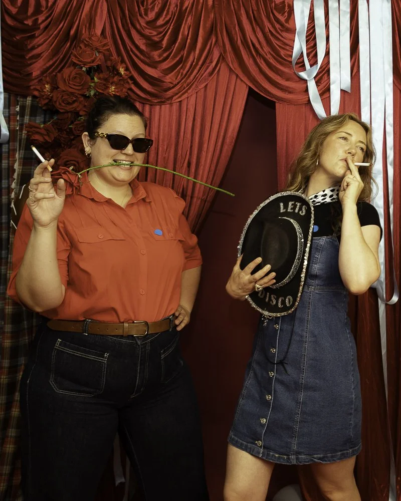 Two women at a party with red curtains and decorations. The woman on the left is wearing sunglasses, a red shirt, and holding a cigarette in one hand and a long green plant in her mouth. The woman on the right is holding a hat that says 'Let's Disco,