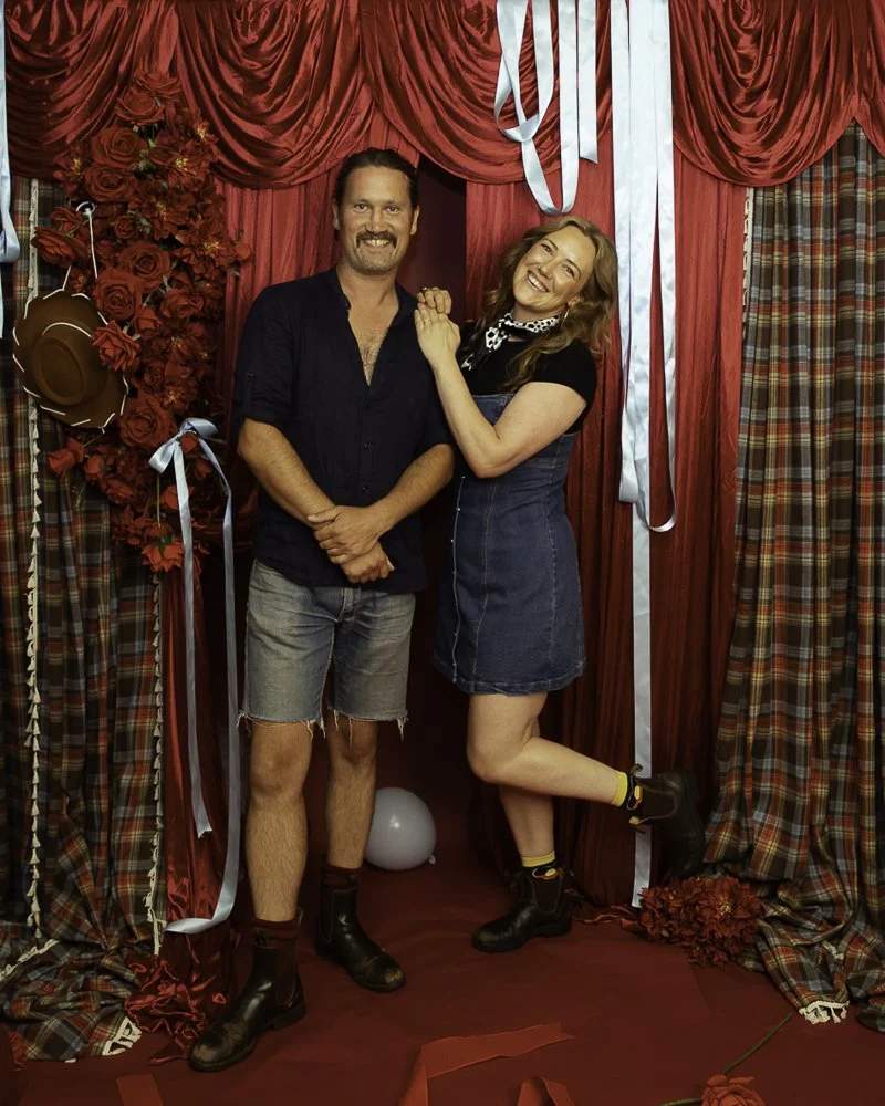 A man and woman standing together in front of red curtains and floral decorations, smiling and holding hands, with the woman leaning on the man's shoulder.