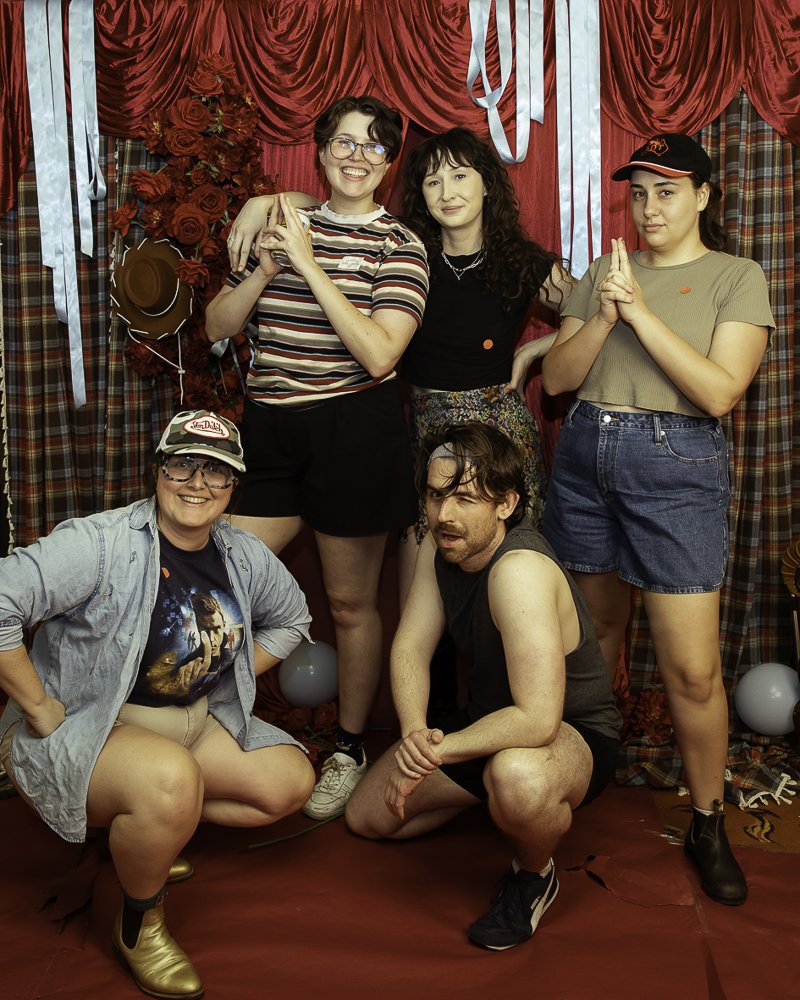Group of five people posing in front of a red and plaid curtain backdrop, with decorations including flowers, balloons, hats, and ribbons.