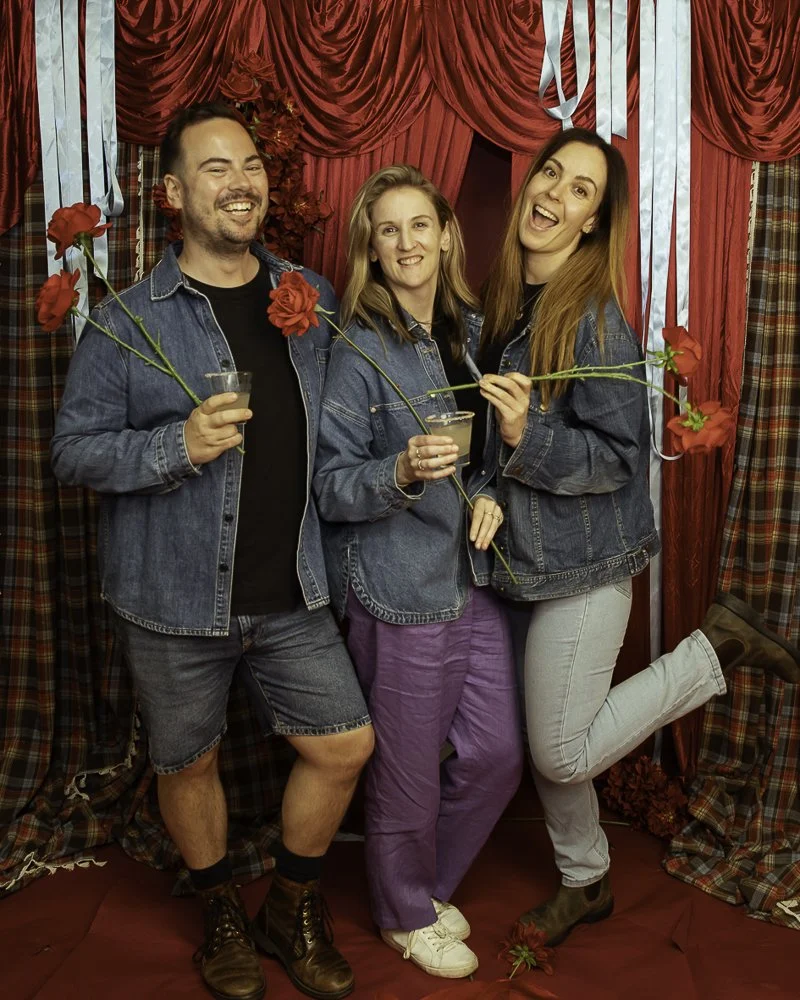 Three friends smiling and holding roses and drinks in front of red curtains at a party or celebration.