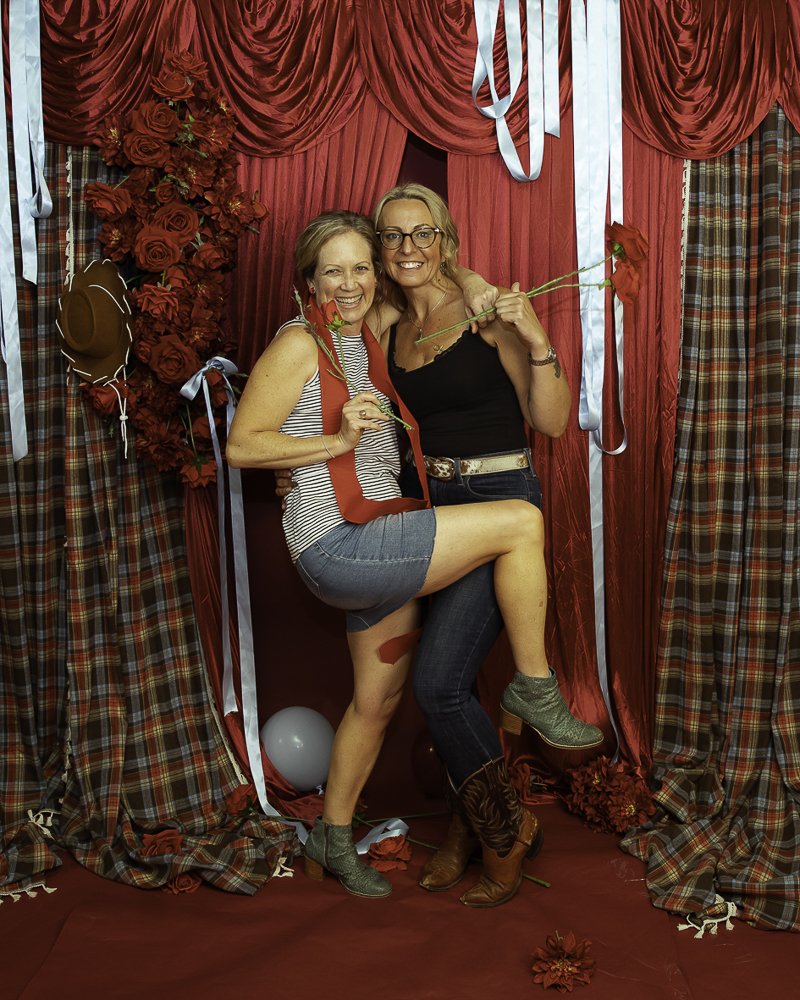 Two women smiling in a celebratory setting decorated with red roses, plaid curtains, and red drapes. One woman is holding a red rose, and the other is holding a flower. One woman is wearing a striped shirt and shorts, and the other is wearing a black