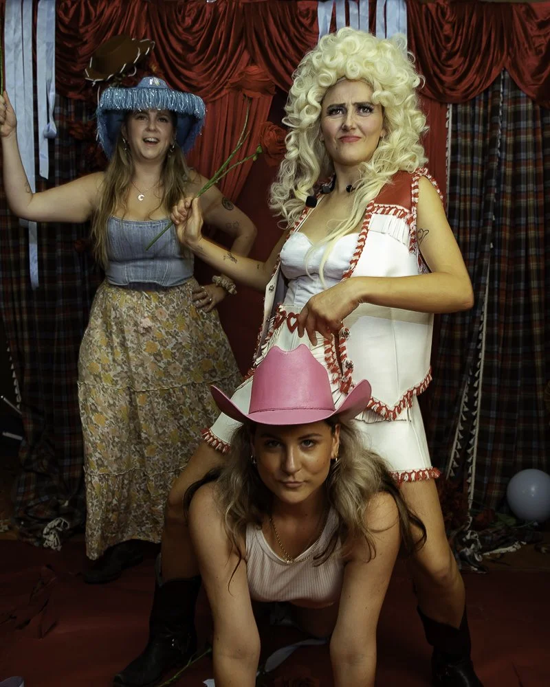 Three women dressed in costumes posing in front of a red curtain. The woman in front is wearing a pink cowboy hat, the woman in the middle has voluminous blonde hair and is dressed in a white and red ruffled outfit, and the woman in the back is weari
