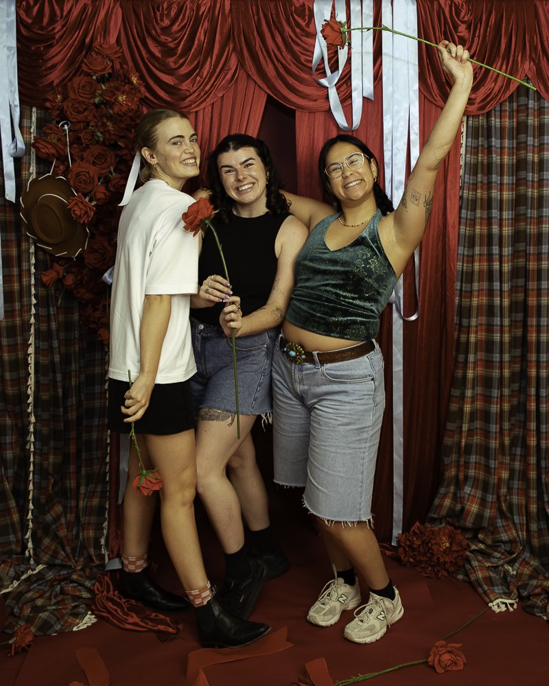 Three women smiling in front of a decorated curtain with red roses and ribbons, holding roses, with one raising a rose in the air.