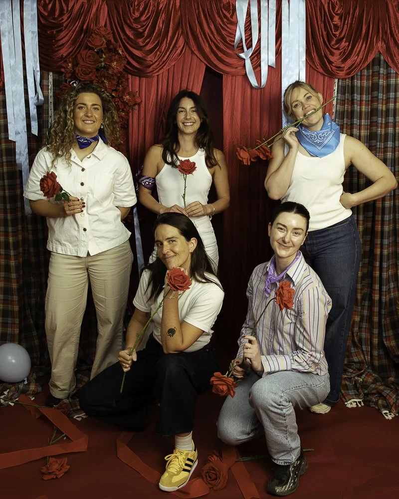 Five women posing in front of a red and plaid curtain background, holding red roses, with red roses and ribbons on the floor and blue ribbons hanging from the ceiling.