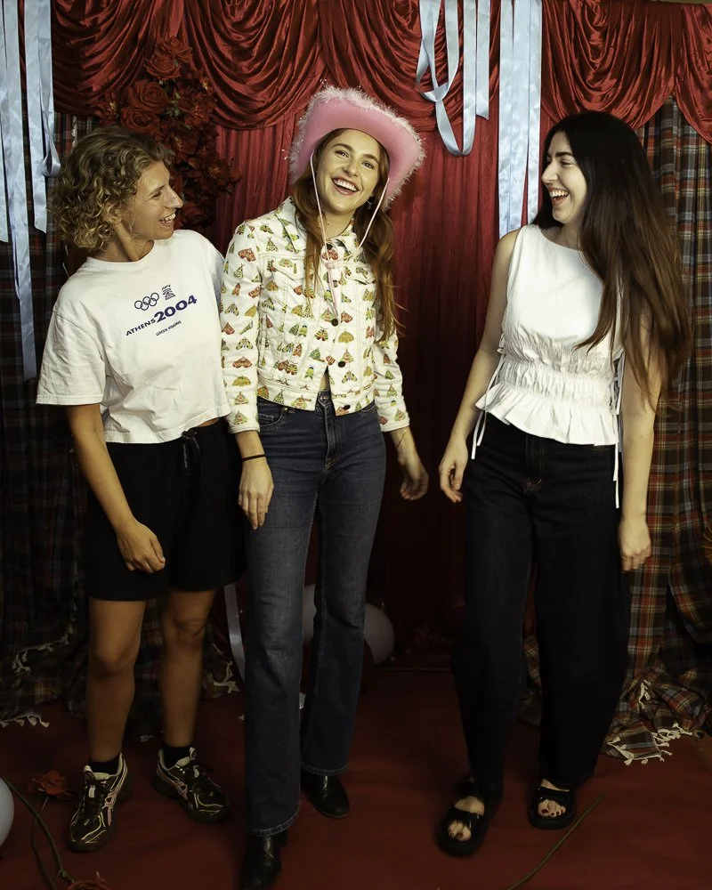 Three women standing in front of red curtains with blue ribbons, smiling and laughing during a celebration. The woman in the middle wears a pink hat and a cardigan with a colorful pattern, while the other two women wear casual outfits with one in a w