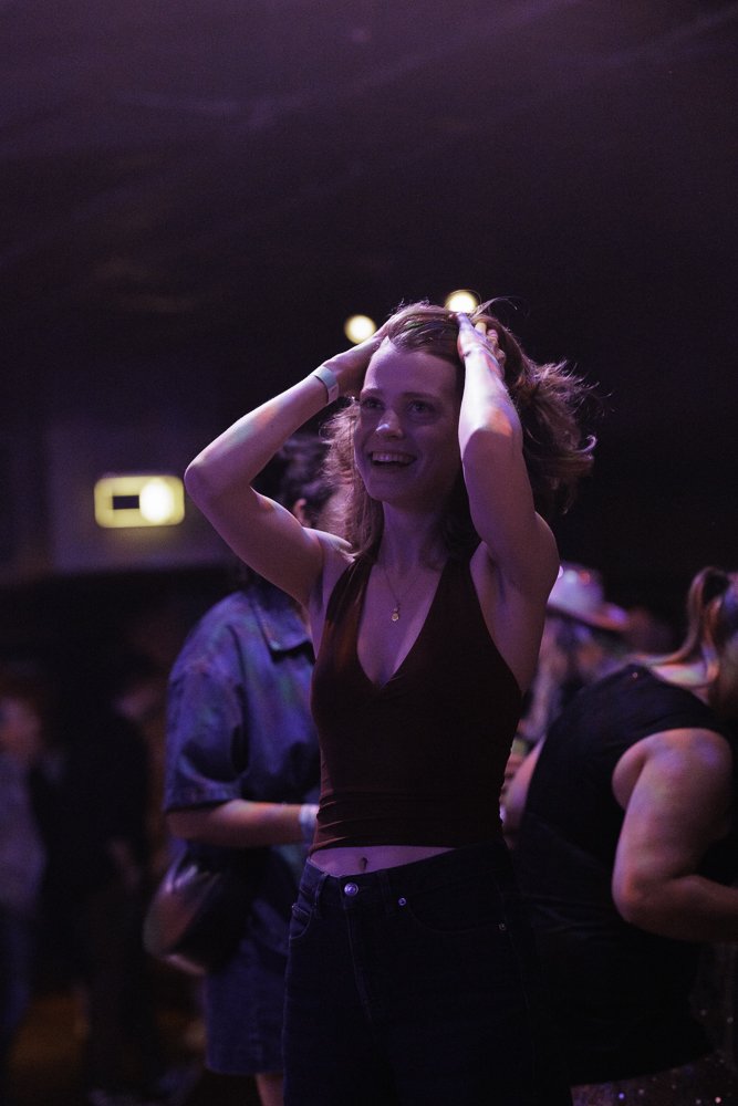 A young woman with light brown hair smiling and holding her hands on her head, standing in a dimly lit indoor setting with purple and pink lighting, surrounded by other people.