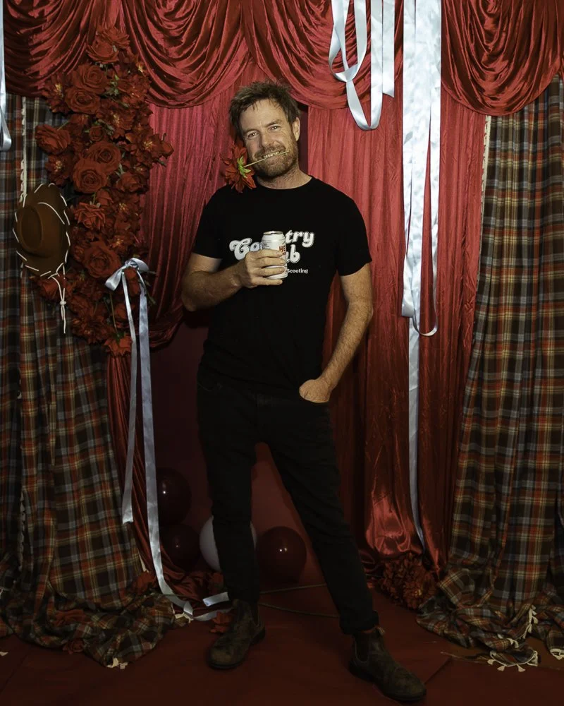 A man with a beard and messy hair standing in front of red and plaid curtains, holding a can of White Claw, with a flower in his mouth, in a decorated setting with balloons and ribbons.