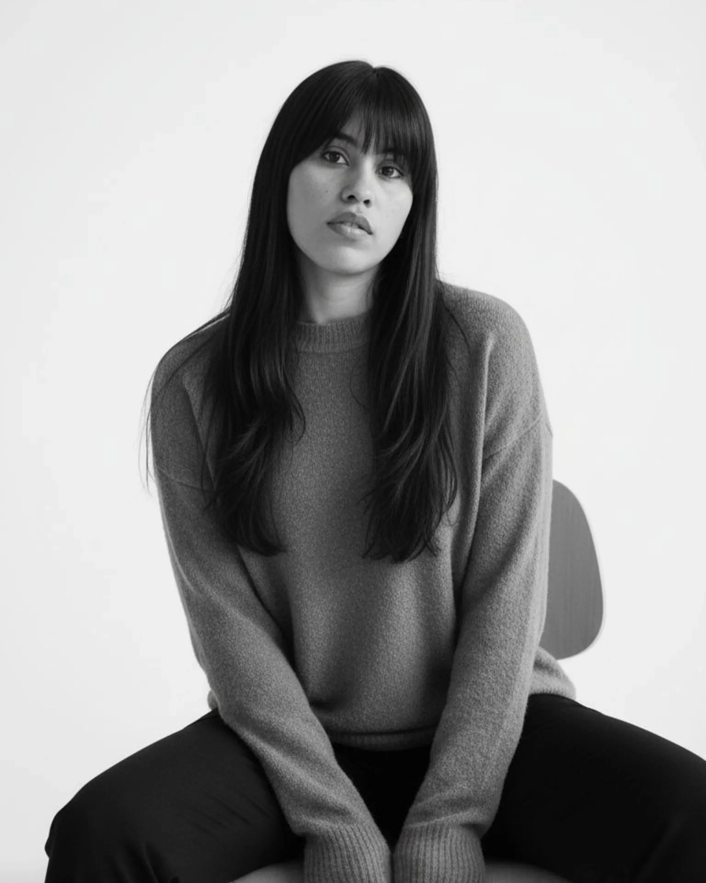 A black and white photo of a woman with long, straight dark hair and bangs, sitting on a chair against a plain background, wearing a cozy sweater and dark pants, looking slightly to her right.