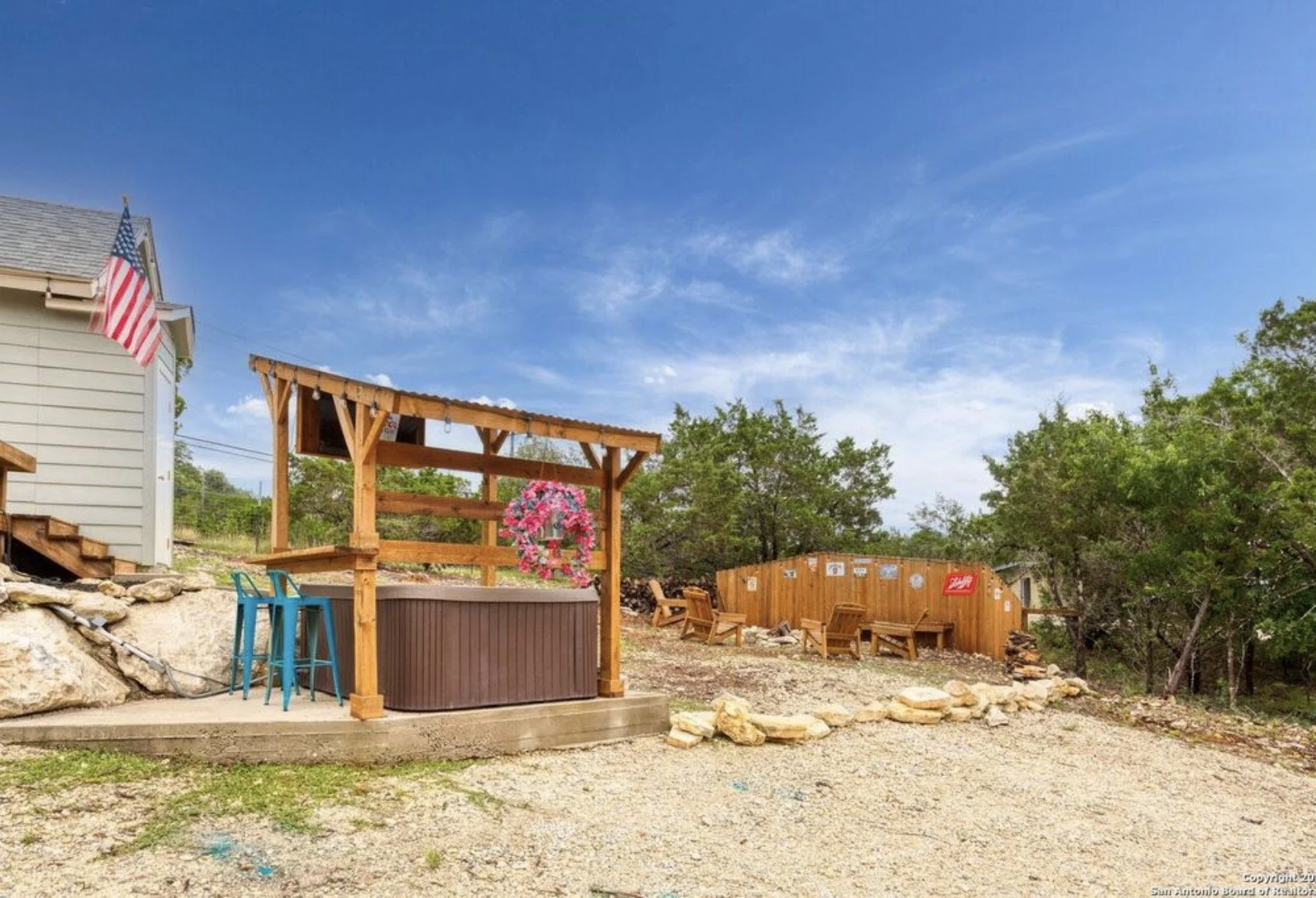 Outdoor backyard area with a hot tub under a wooden pergola, decorated with a pink floral wreath, and several blue chairs nearby. A gravel ground with rocks and a wooden privacy fence with signs. There are trees and a clear blue sky in the background.