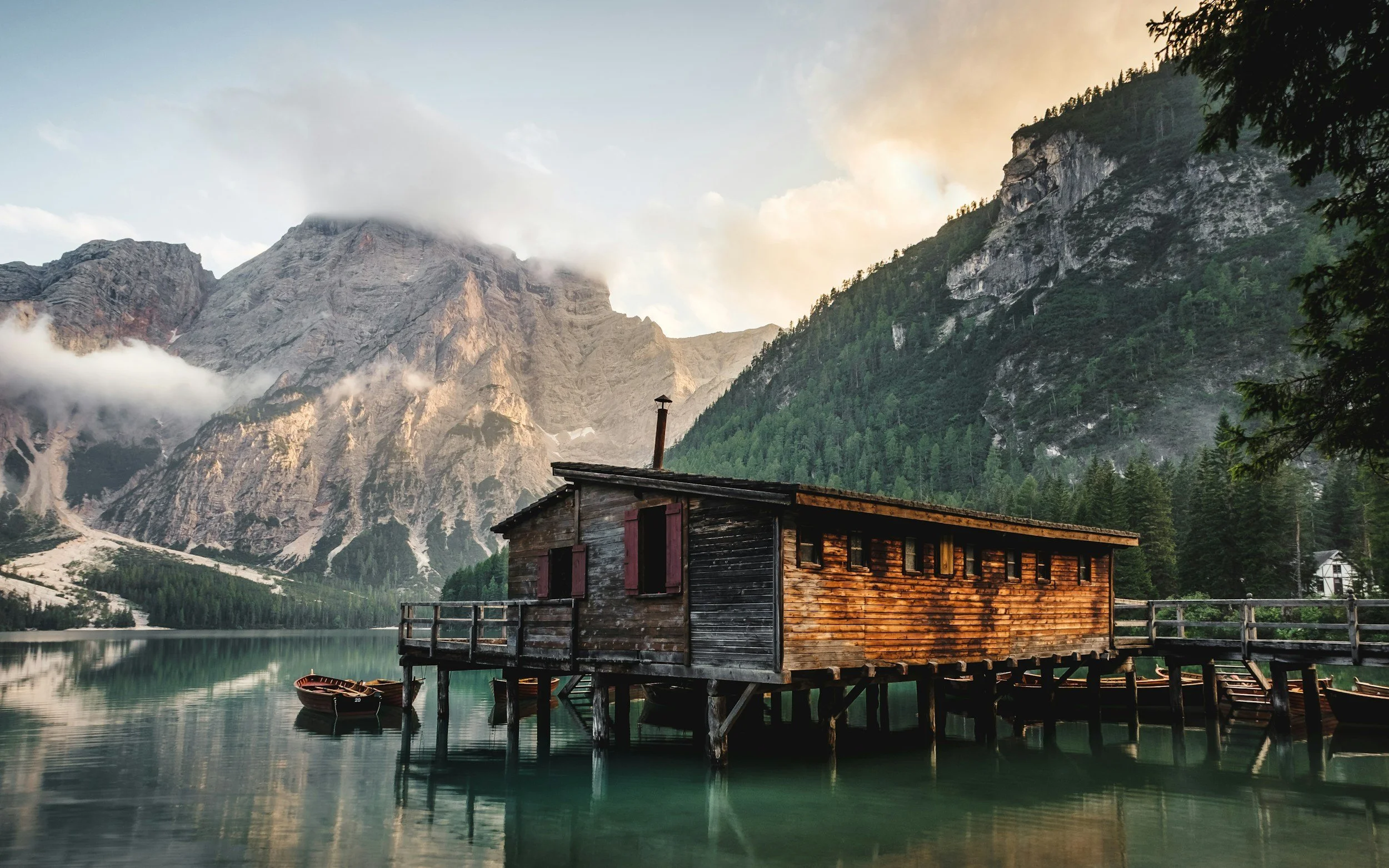 A rustic wooden cabin built on stilts over a calm lake with mountains and a forest in the background.