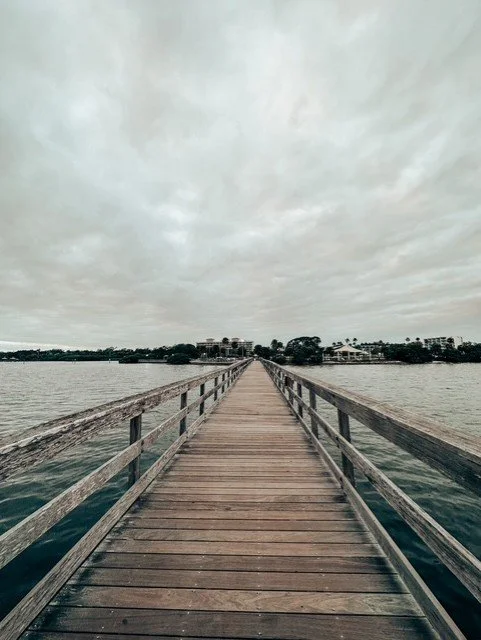 A wooden pier extending over water towards a cloudy sky with buildings and trees in the distance.