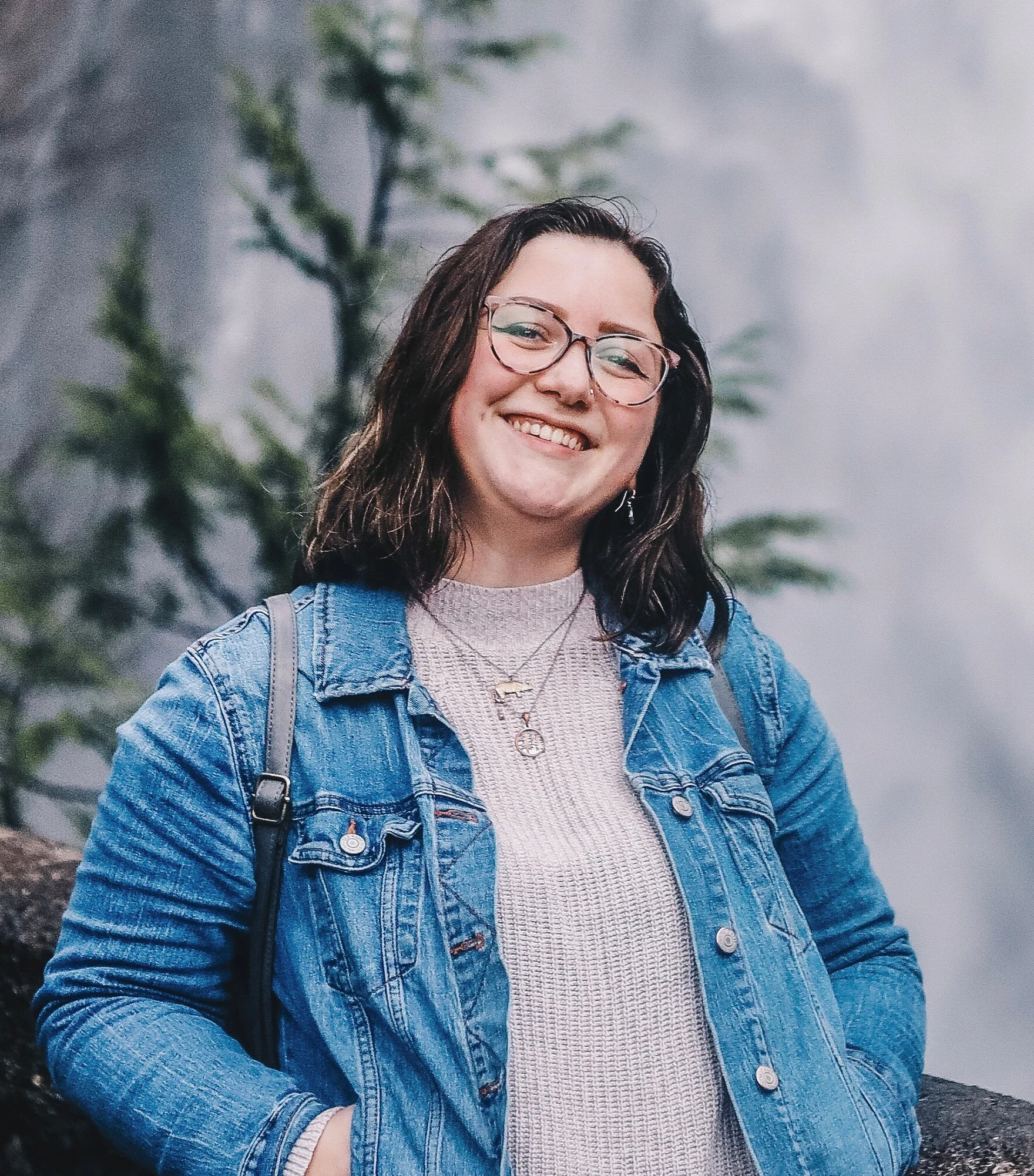 A smiling woman with glasses, wearing a denim jacket and layered necklaces, outdoors with a misty background of trees.