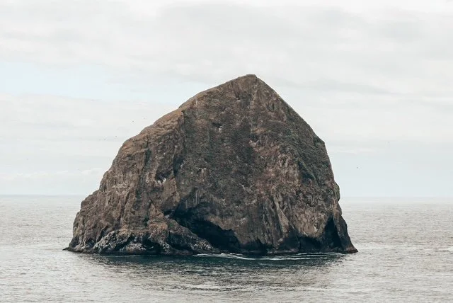 Large ocean rock formation with a steep, angular shape, surrounded by water under a cloudy sky.