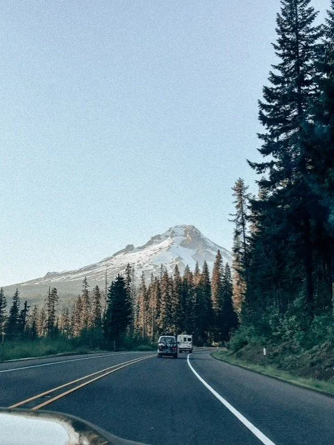 A mountain with snow on its peak, trees on either side of a winding road, and vehicles driving towards the mountain.