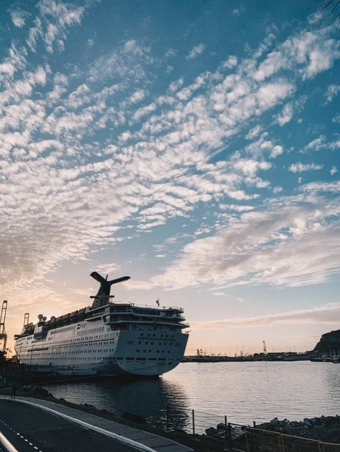 A large cruise ship docked at a port, with a partly cloudy sky overhead and calm water in the foreground.