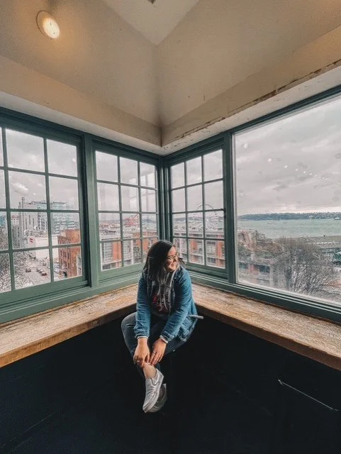 Woman sitting by large corner windows in a high-rise building, overlooking cityscape and water.