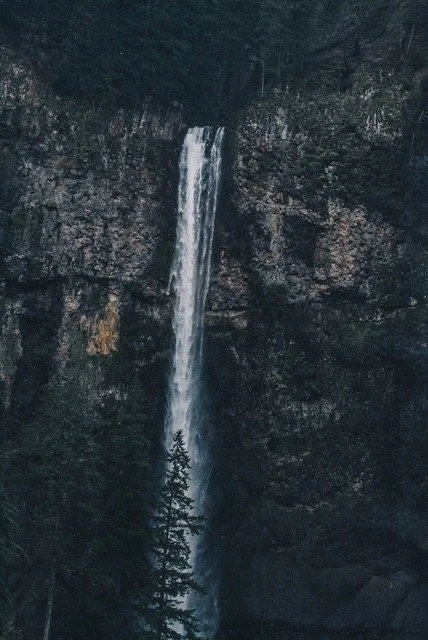 A tall waterfall cascading down a rocky cliff into a dark forested area below.