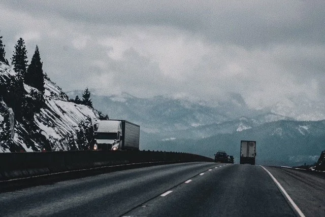 Highway with vehicles traveling through mountainous, snowy landscape under cloudy sky.
