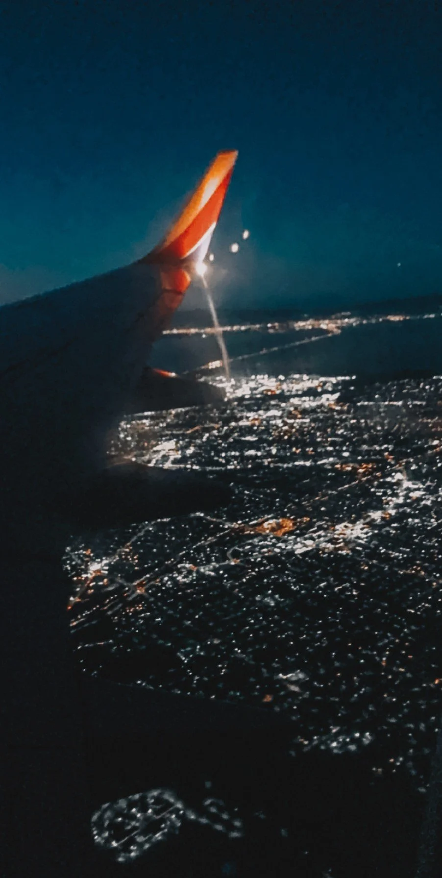 Nighttime aerial view from an airplane window showing city lights below and the airplane wing with a visible logo.