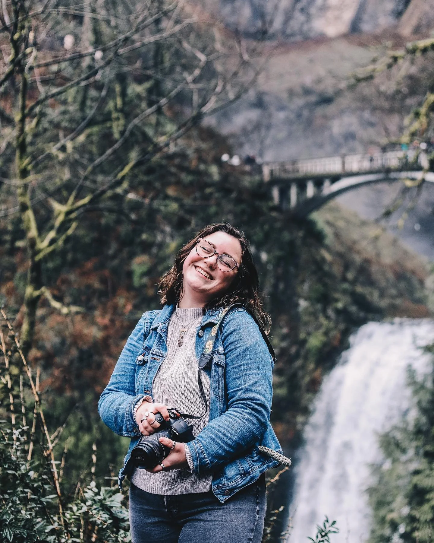 A woman standing outdoors near a waterfall, holding a camera, smiling, wearing glasses, a denim jacket, and a beige sweater.