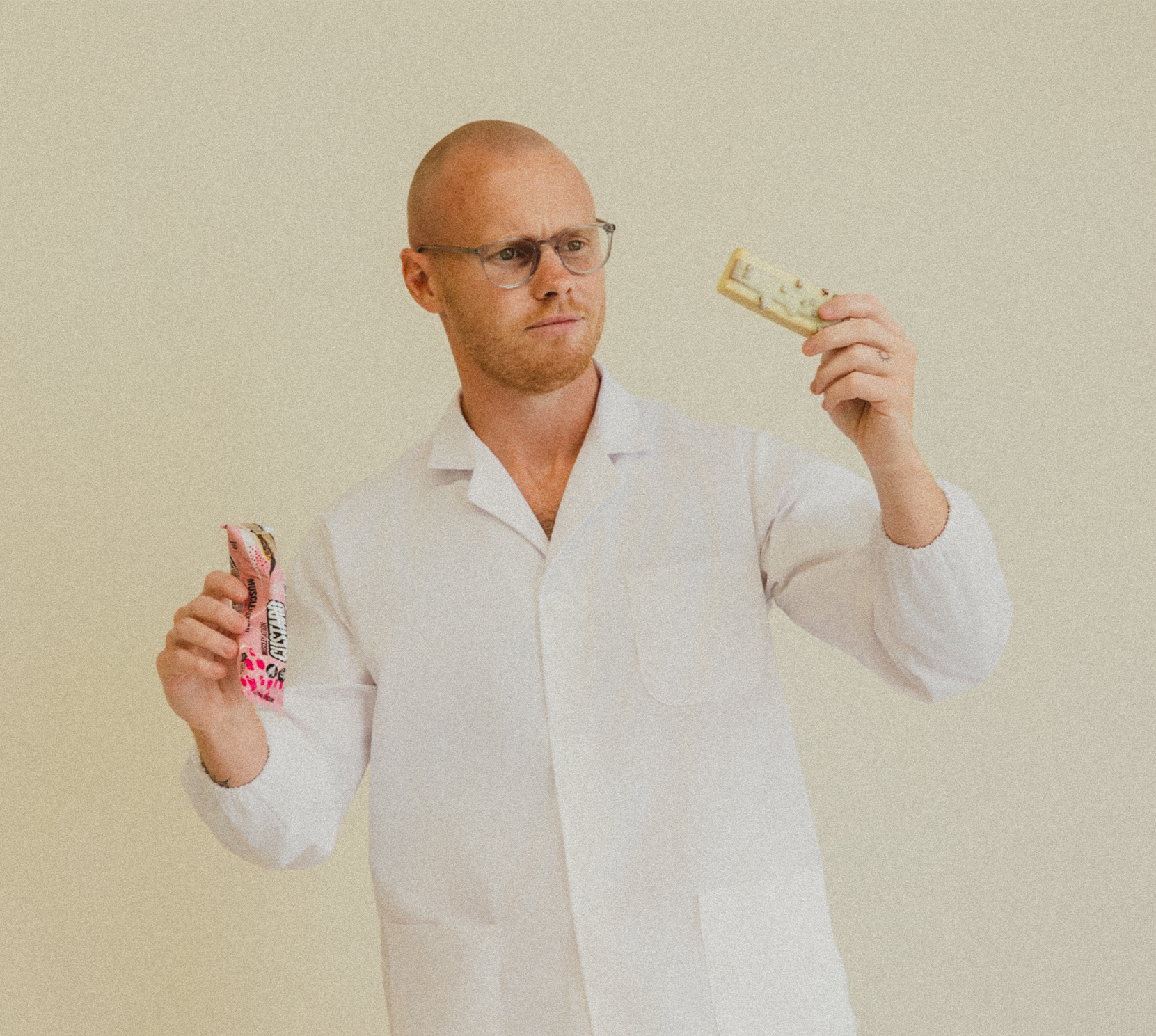 Lachy Oliver wearing a lab coat and glasses holding a protein bar looking at it confused