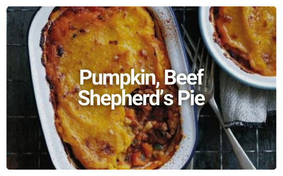 A dish of pumpkin and beef shepherd's pie topped with mashed potatoes in a white oval baking dish, with a smaller serving on a plate with a spoon, set on a dark surface with a gray napkin.