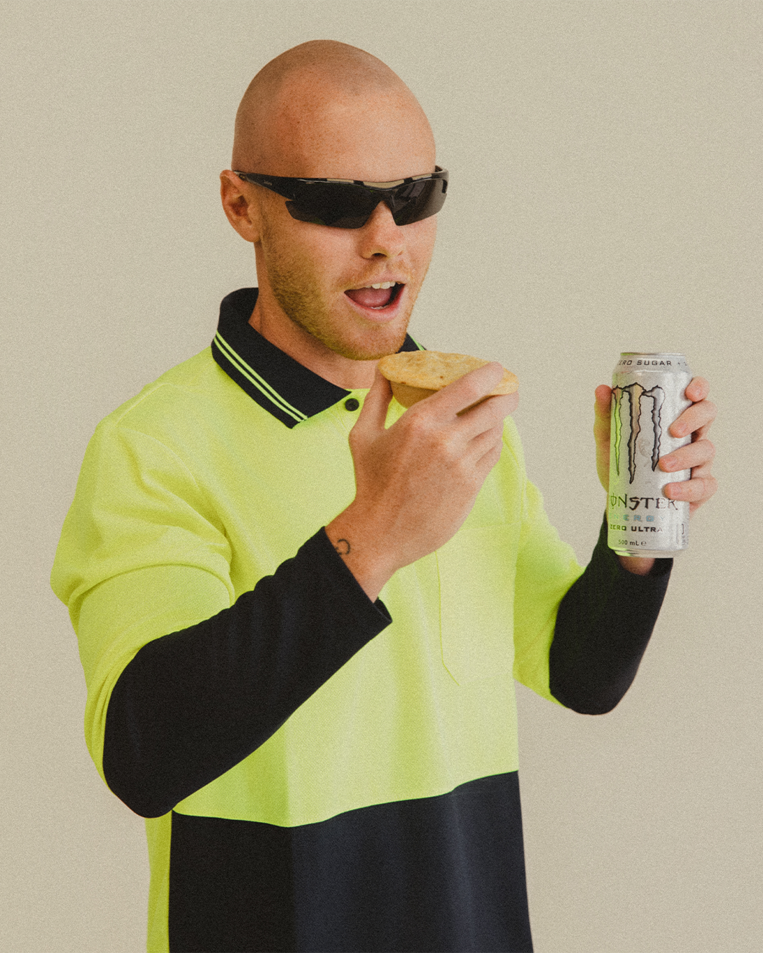 Lachy Oliver wearing a neon yellow tradesman shirt and visor sunglasses holding a white monster can and a pie to his mouth