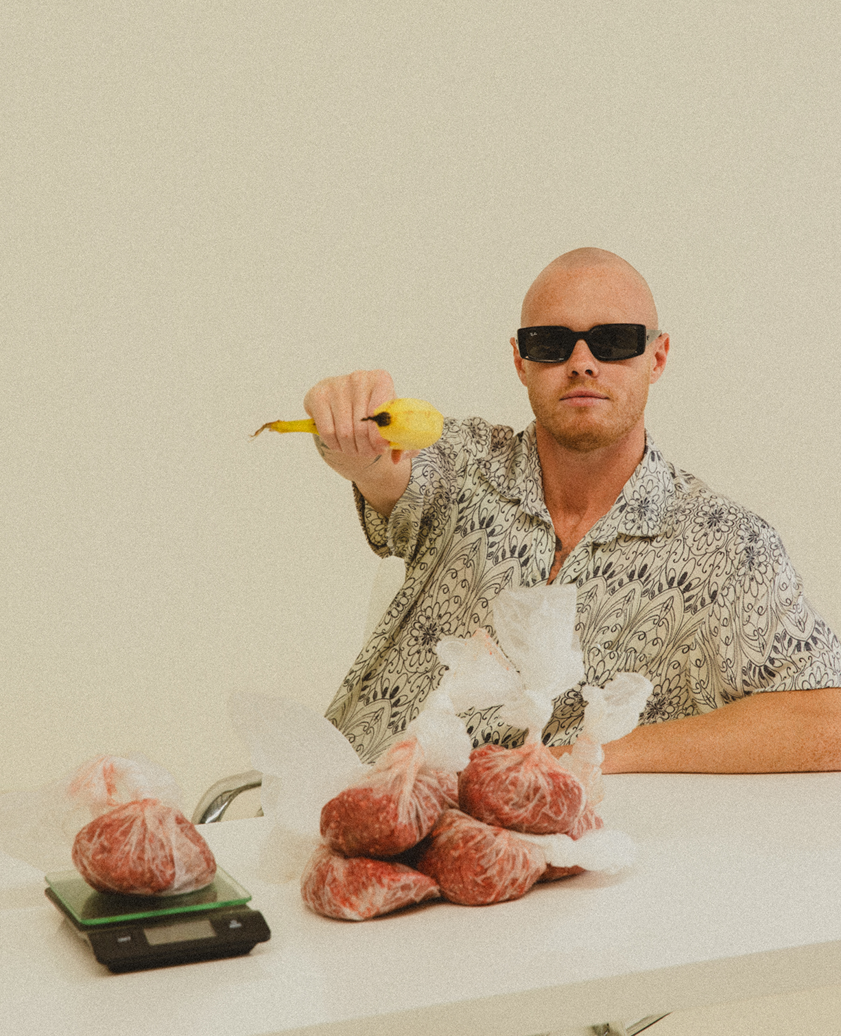 Lachy Oliver wearing a patterned shirt and black sunglasses pointing a banana at the camera with mince weighed up on a table in plastic bags