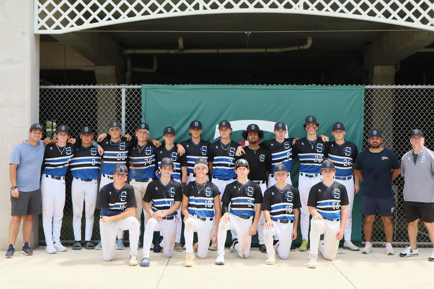 Showtime Select 15U with coaches posing for team photo in front of a chain-link fence and green backdrop, wearing matching black and blue uniforms and caps.