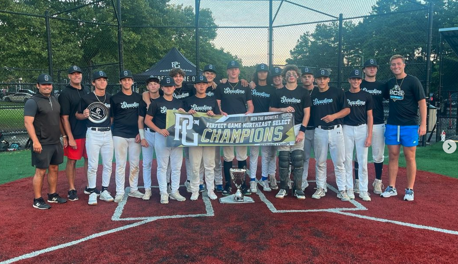 A youth baseball team in black shirts and white pants celebrating on a baseball field after winning a championship, with a trophy and a banner reading 'Perfect Game Northeast Select Champions.' The team is flanked by two coaches or staff members.