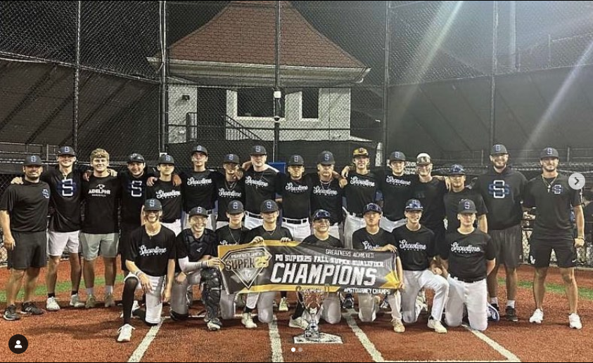 Baseball team posing on field at night holding a Champions banner and trophy, wearing black and white uniforms and caps, with a chain-link fence and buildings in background.