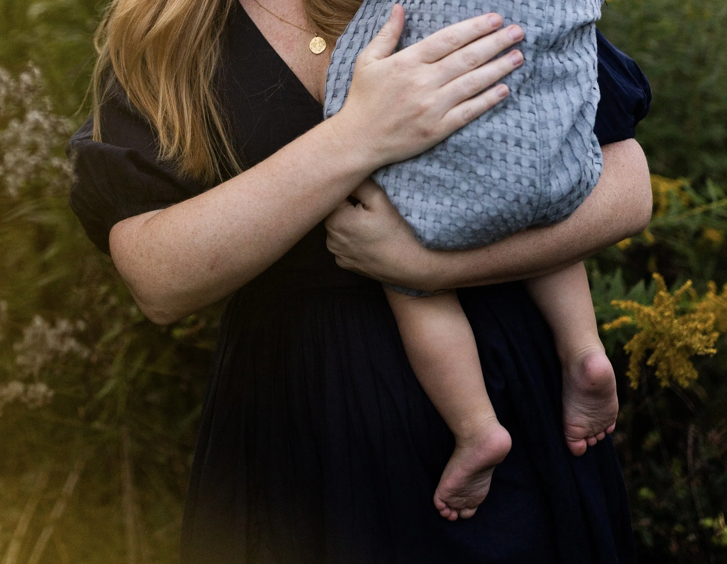 A person holding a child outdoors, with the person's hand supporting the child's back and arm, in a natural setting with greenery in the background.