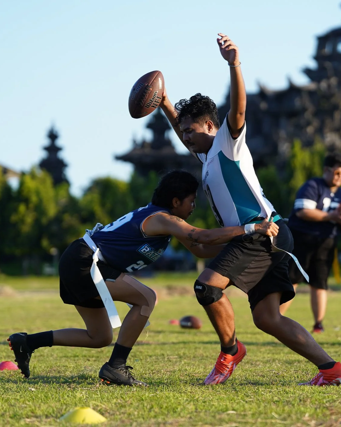 Two young men playing Australian football on a field, with one man holding a football and the other tackling him. There are trees and traditional buildings in the background.