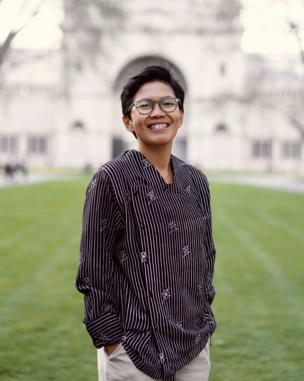 A young person with short dark hair, glasses, and braces standing outdoors on a grassy area with a large, historic building in the background.