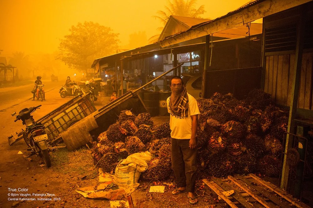 A man wearing a scarf mask stands next to a large pile of palm fruit for oil extraction outside a shop, with a fallen motorbike and a leaning pole in the background, amid a hazy orange sky caused by wildfire smoke in Palangka Raya, Indonesia, in 2015.