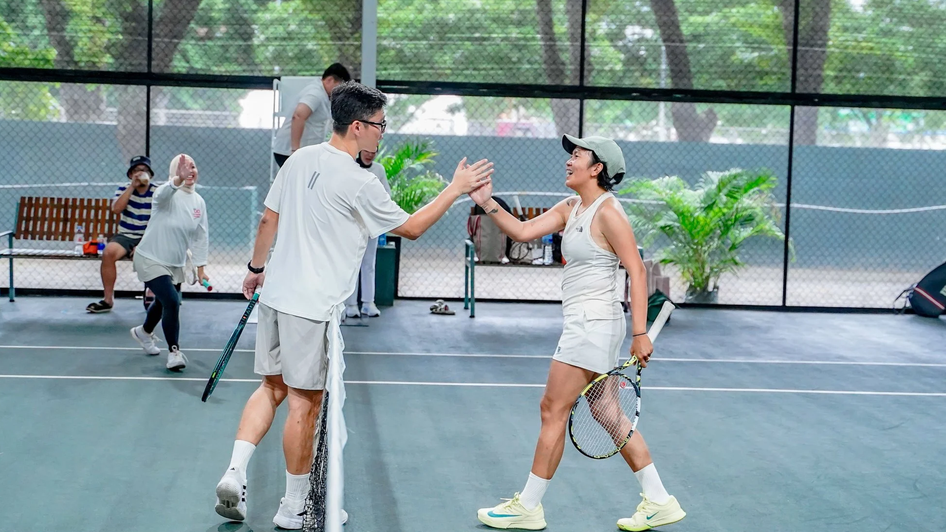 Two people on a tennis court, high-fiving after a match or practice. The man is wearing glasses, a white athletic shirt, and shorts, holding a tennis racket. The woman is wearing a white tank top, shorts, a cap, and tennis shoes, holding a tennis racket. In the background, other people are playing or sitting on benches. The court is enclosed with a fence, and there are trees outside.