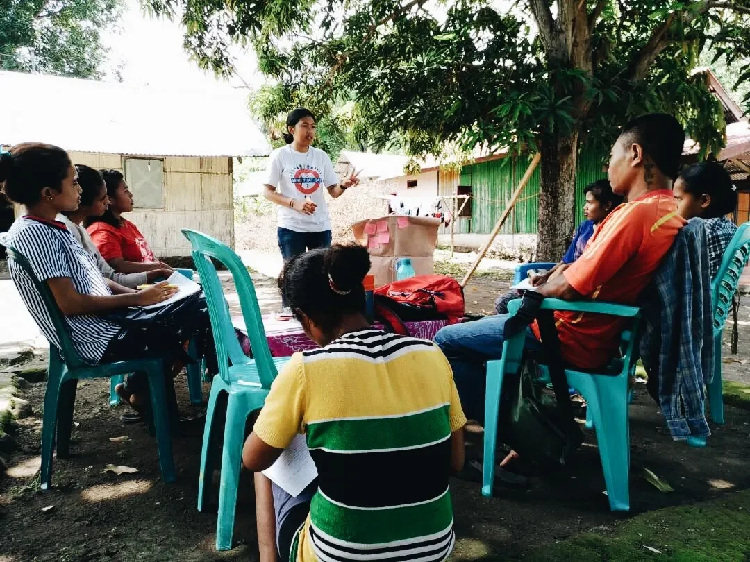 A woman standing in front of a group of women, giving a presentation outdoors under a tree with small houses or structures in the background.