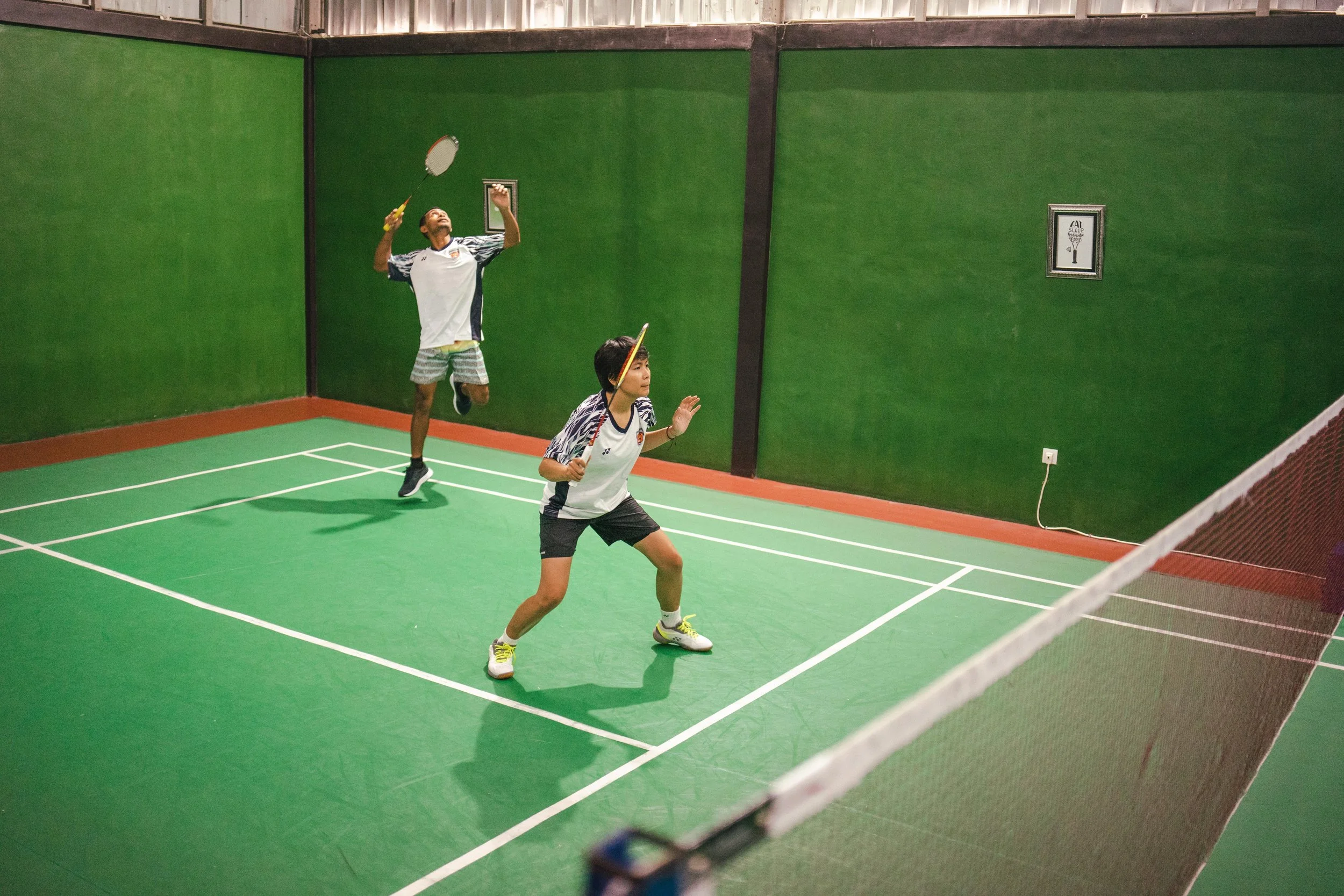 Two people playing badminton on a green court with white lines, enclosed by green walls and a red border. One person is jumping to hit the shuttlecock while the other is preparing to hit back.