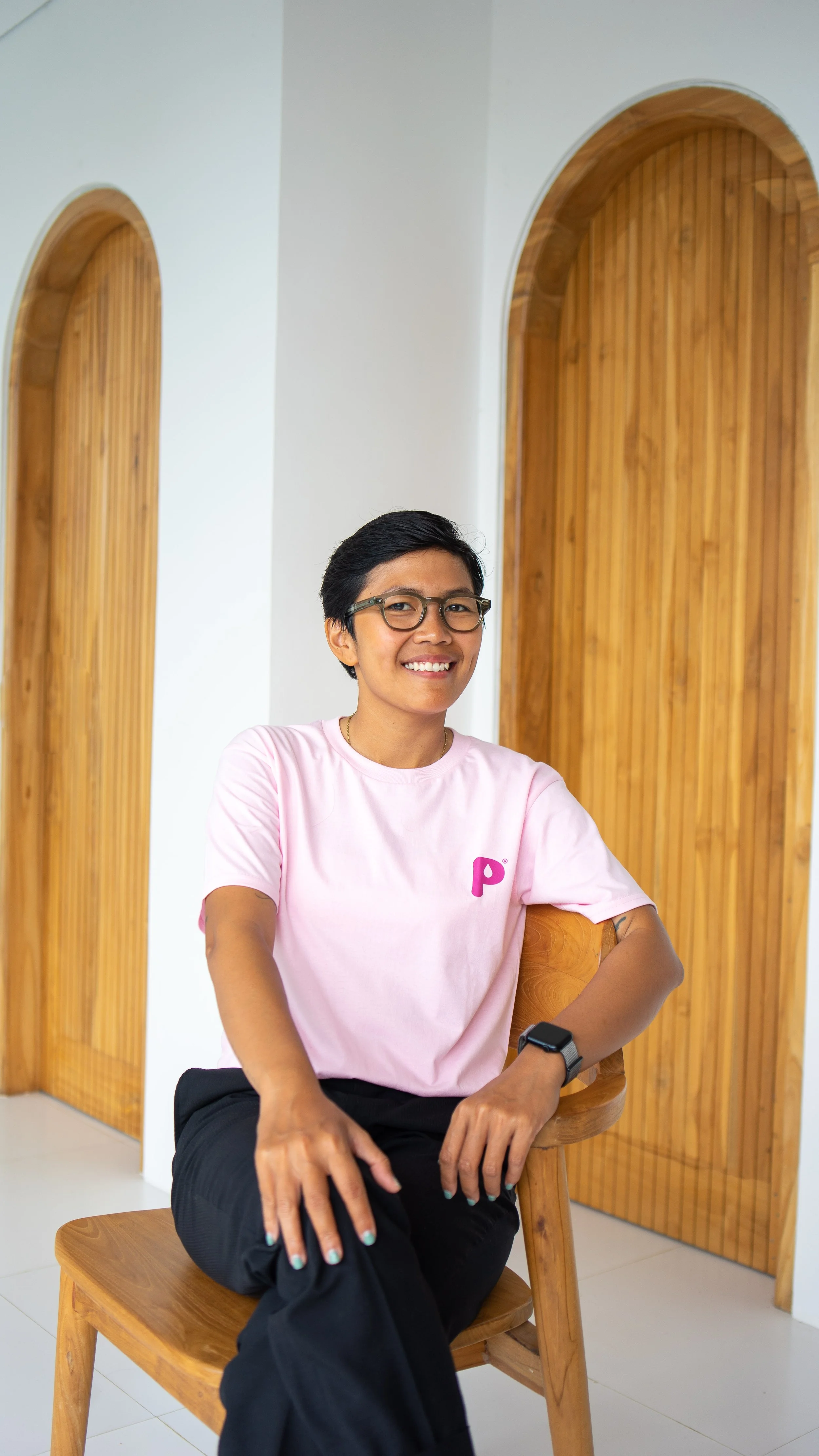 A woman with short black hair, glasses, and a pink t-shirt with a purple 'P' logo, sitting on a wooden chair with a wooden back in front of a white wall with wooden arched doorways, smiling at the camera.