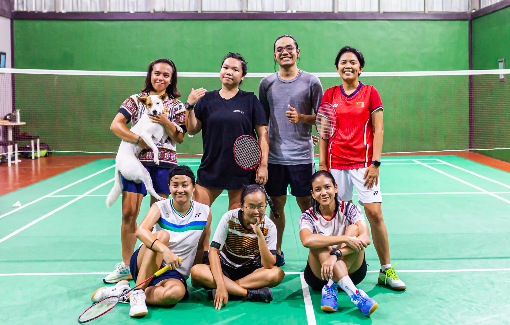 Group of eight women and one dog on a badminton court, holding rackets and smiling, with a green wall and badminton net in the background.