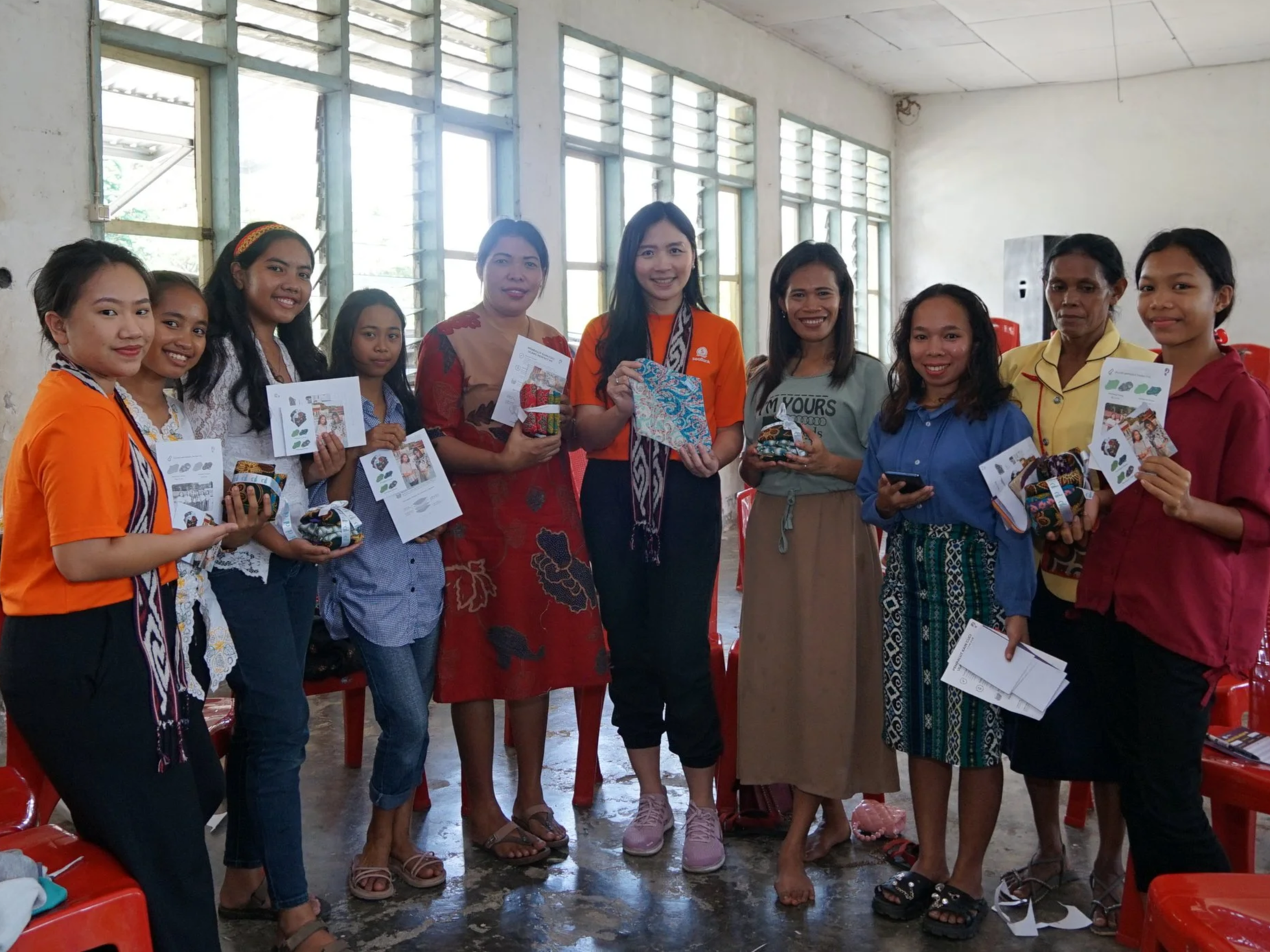 Group of women and girls standing together indoors, holding small wrapped gifts and flyers, smiling at the camera.