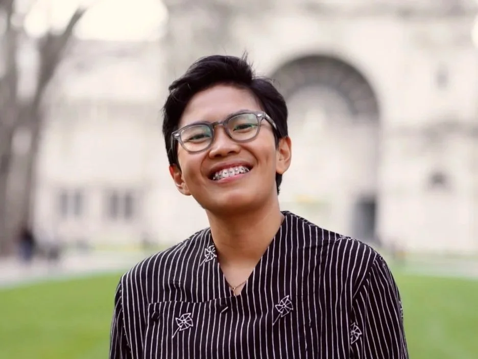 A young man with glasses and braces smiling outdoors in front of a blurred building and grass.
