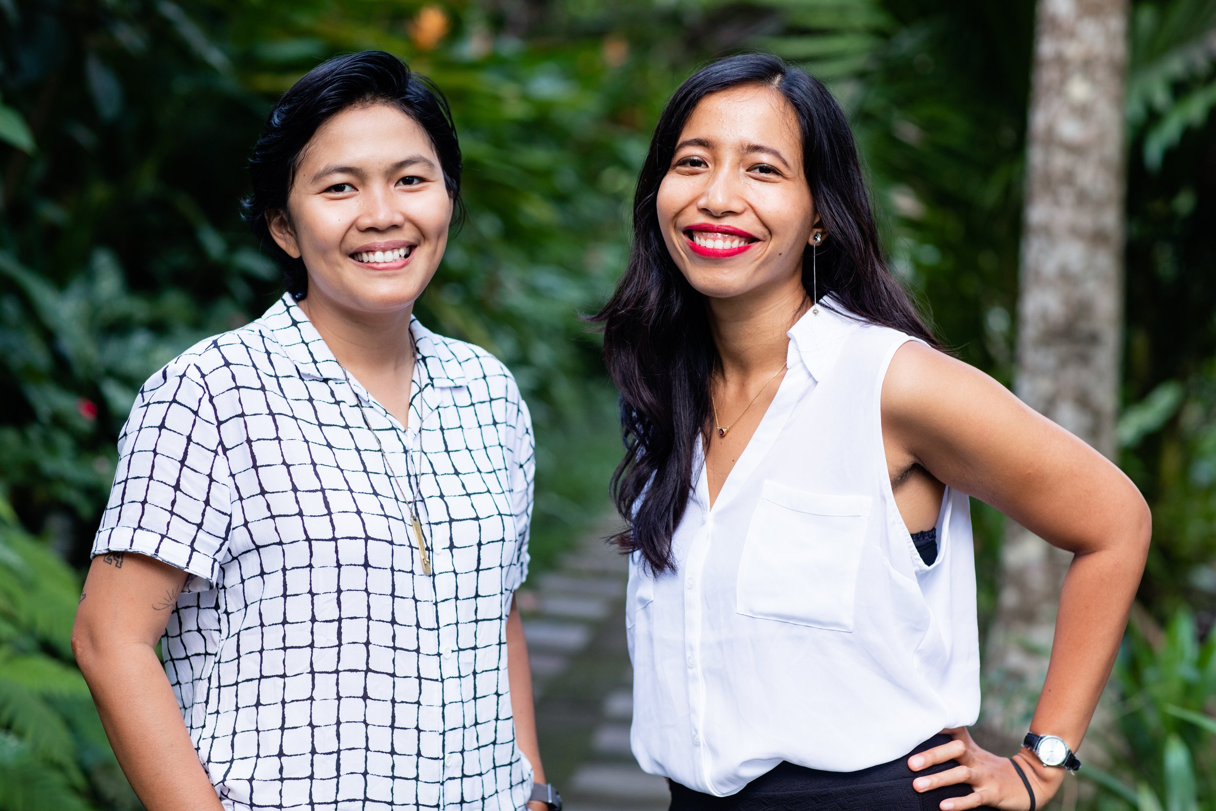 Two women standing outdoors in a green, tropical environment, smiling at the camera.