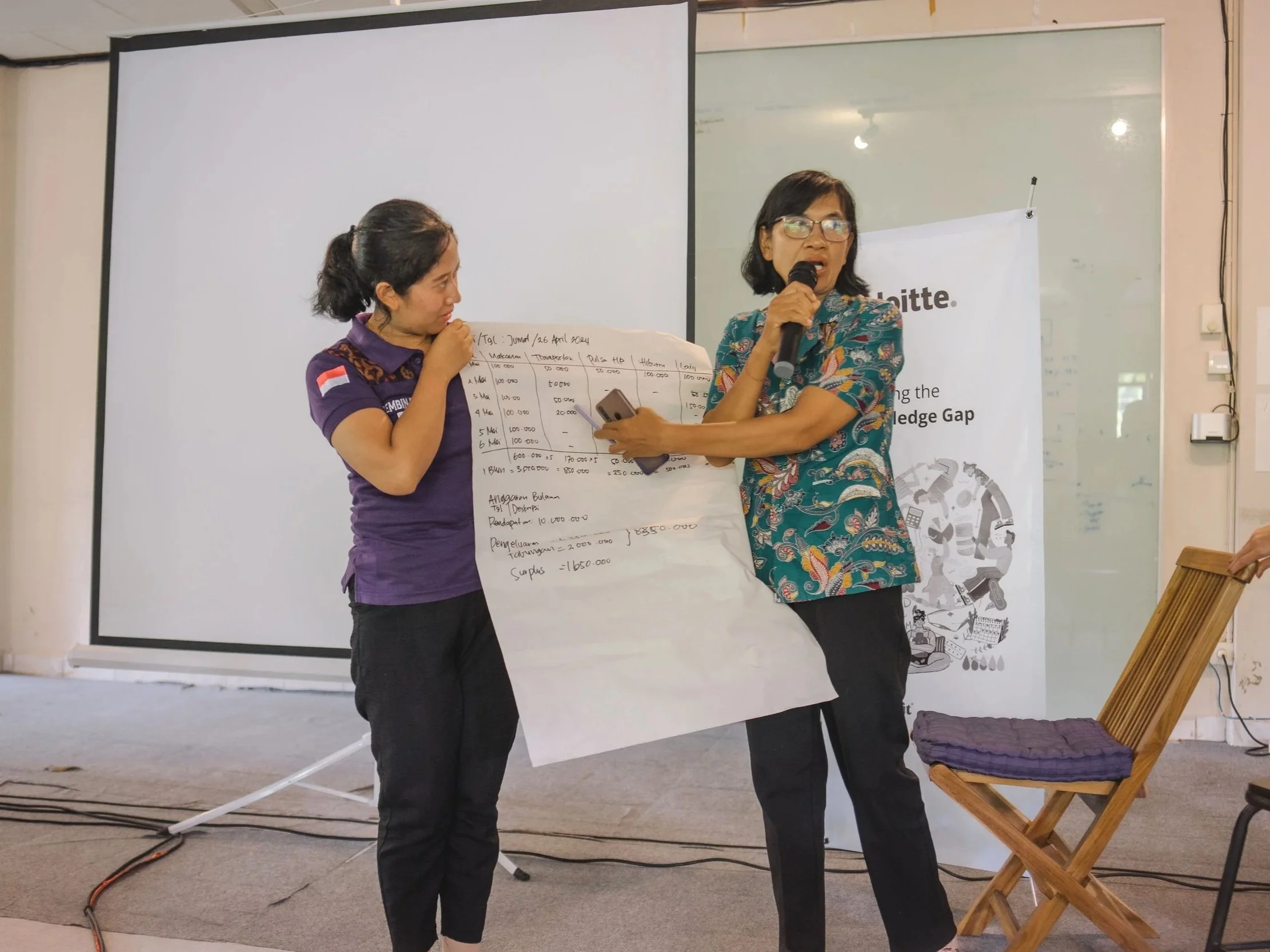 Two women presenting on stage with a large sheet of paper containing handwritten notes or data. One woman is holding a microphone and pointing at the paper, while the other looks on, holding the paper. A banner and a chair are visible in the background.
