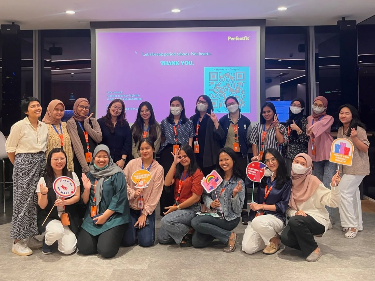 Group of women at an indoor event, some wearing masks, holding signs and making peace signs, with a large screen in the background displaying a purple slide with text and a QR code.
