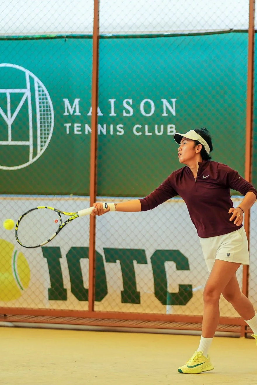 A woman playing tennis on an outdoor court at Maison Tennis Club, hitting a tennis ball with a racquet.