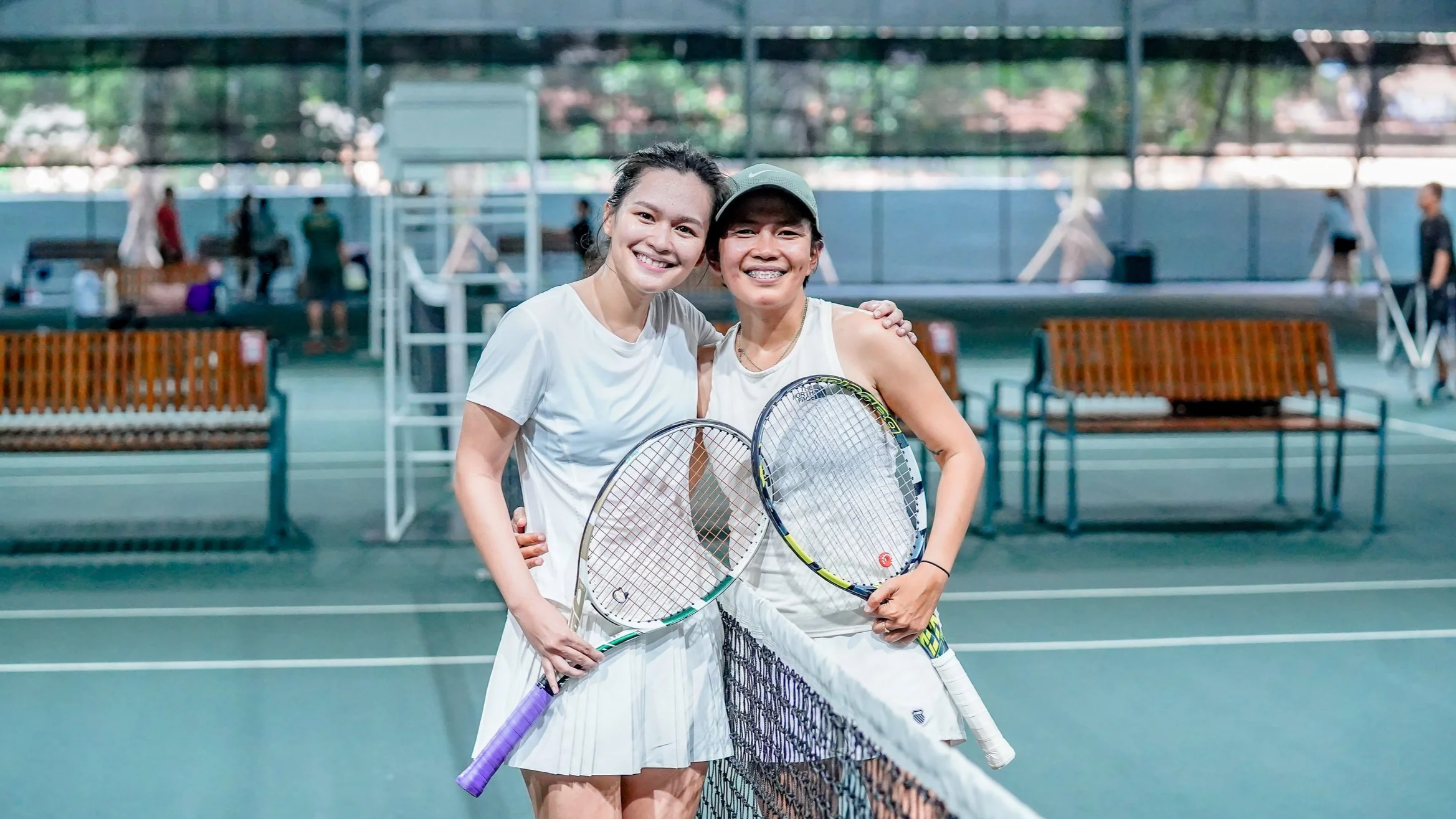 Two women standing on an indoor tennis court holding tennis rackets, smiling for a selfie, with other players and tennis equipment in the background.